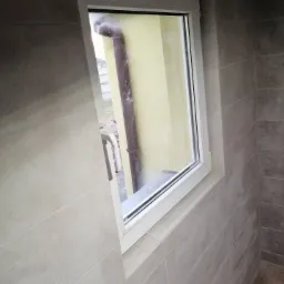 Szare płytki ceramiczne ułożone poziomo na ścianie i podłodze, z białym oknem PCV w rogu pomieszczenia, widok na rynnę za oknem.