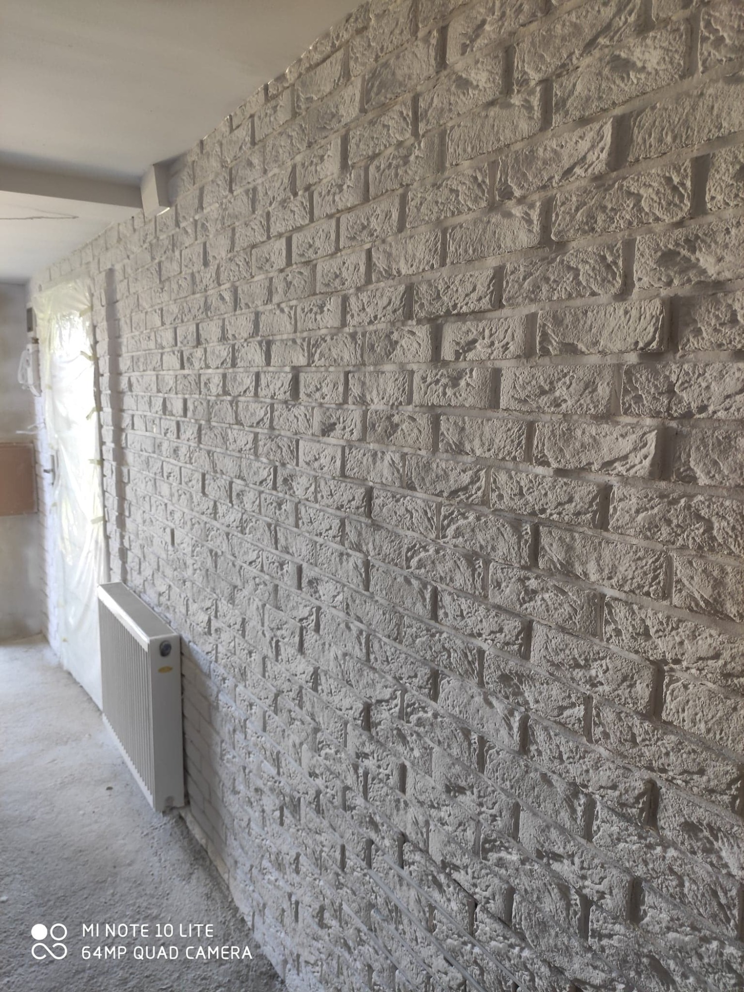 Ściana wykończona dekoracyjną cegłą w odcieniach szarości, z grzejnikiem przy ścianie i widocznym zabezpieczonym folią ościeżem drzwiowym w tle.