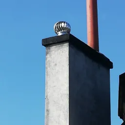 Komin z zamontowaną metalową nasadą obrotową, widoczny z dołu na tle błękitnego nieba, z dodatkową rurą kominową w tle.