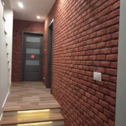 Korytarz z oświetlonymi schodami, ścianą wykończoną czerwoną cegłą i szarą, nowoczesną stolarką drzwiową.