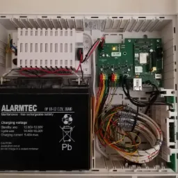 Otwarta skrzynka centrali alarmowej z akumulatorem ALARMTEC, płytką drukowaną JABLOTRON JA-107K oraz wiązką kolorowych kabli, widok z góry.