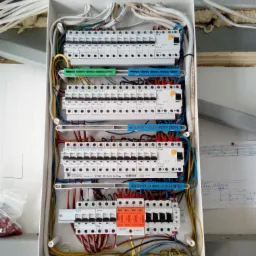 Rozdzielnica elektryczna w białej obudowie, widoczne liczne bezpieczniki EATON, przewody w różnych kolorach, listwy zaciskowe w odcieniach zieleni i niebieskiego, schemat instalacji elektrycznej...