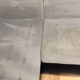 Fragment szarej, narożnej kanapy z widoczną różnicą w czystości tapicerki po zabiegu czyszczenia, z wyraźnymi plamami na nieczyszczonej części.