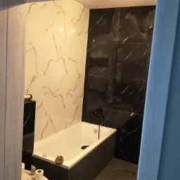 Nowoczesna łazienka z biało-czarnymi marmurkowymi płytkami, białą wanną obudowaną czarnym kamieniem, oraz toaletą z akcesoriami, widziana zza framugi drzwi.