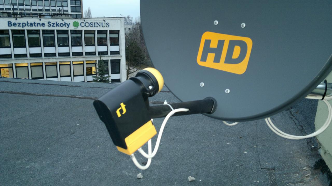 Szara antena satelitarna HD z czarno-żółtym konwerterem unicable na dachu budynku z napisem 'Bezpłatne Szkoły Cosinus' w tle.