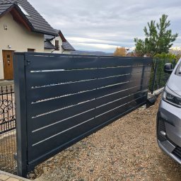 Krystian Zawodny Kris Haus - Nowoczesna, przesuwna brama wjazdowa z poziomymi panelami w kolorze antracytowym, częściowo otwarta, na tle domu jednorodzinnego i żwirowego podjazdu.