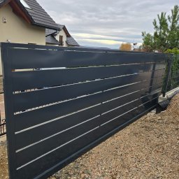 Krystian Zawodny Kris Haus - Nowoczesna, przesuwna brama wjazdowa z poziomymi panelami w kolorze antracytowym na tle domu jednorodzinnego i pochmurnego nieba. Widoczne detale konstrukcyjne.