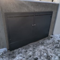Krystian Zawodny Kris Haus - Dwu-skrzydłowa, grafitowa brama garażowa wbudowana w betonowy mur, z widocznymi zawiasami i zamkiem, na tle ośnieżonego podłoża.