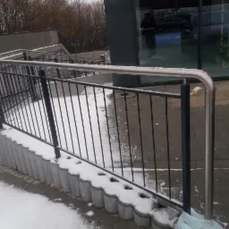 Metalowa balustrada schodowa z elementami malowanymi proszkowo, z widocznym śniegiem na stopniach i otoczeniu budynku.