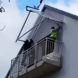 Montaż zadaszenia balkonu z poliwęglanu przez dwóch pracowników na balkonie z metalową barierką, widok z dołu na tle pochmurnego nieba.