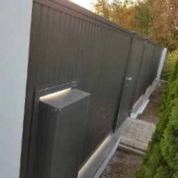 Nowoczesna, przesuwna brama wjazdowa z metalowymi panelami w kolorze antracytowym, z wbudowanym oświetleniem LED, widoczna z boku, z fragmentem ścieżki z kostki brukowej i zielenią w tle.