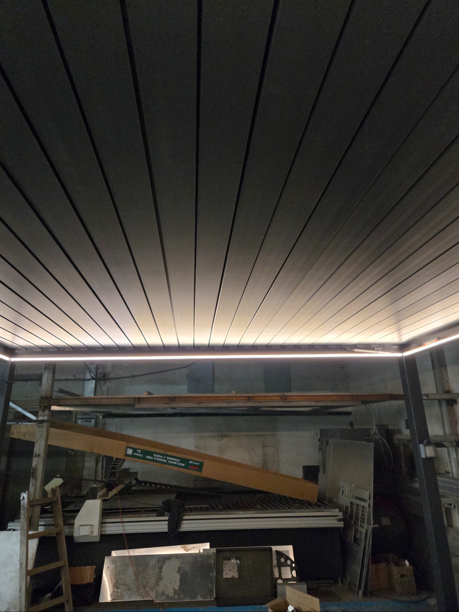 Szare lamele sufitowe z oświetleniem LED wzdłuż krawędzi, widok z dołu, z elementami konstrukcyjnymi i narzędziami w tle. Ukończona instalacja.