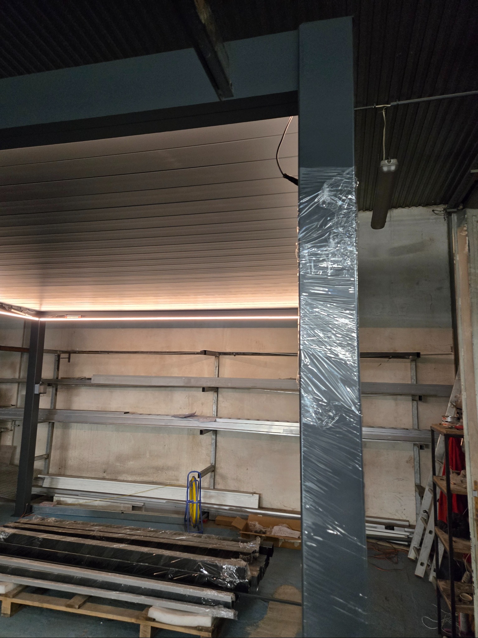 Element konstrukcyjny pergoli, zabezpieczony folią, w hali produkcyjnej. Widoczne profile i regały z materiałami. Oświetlenie LED w tle.