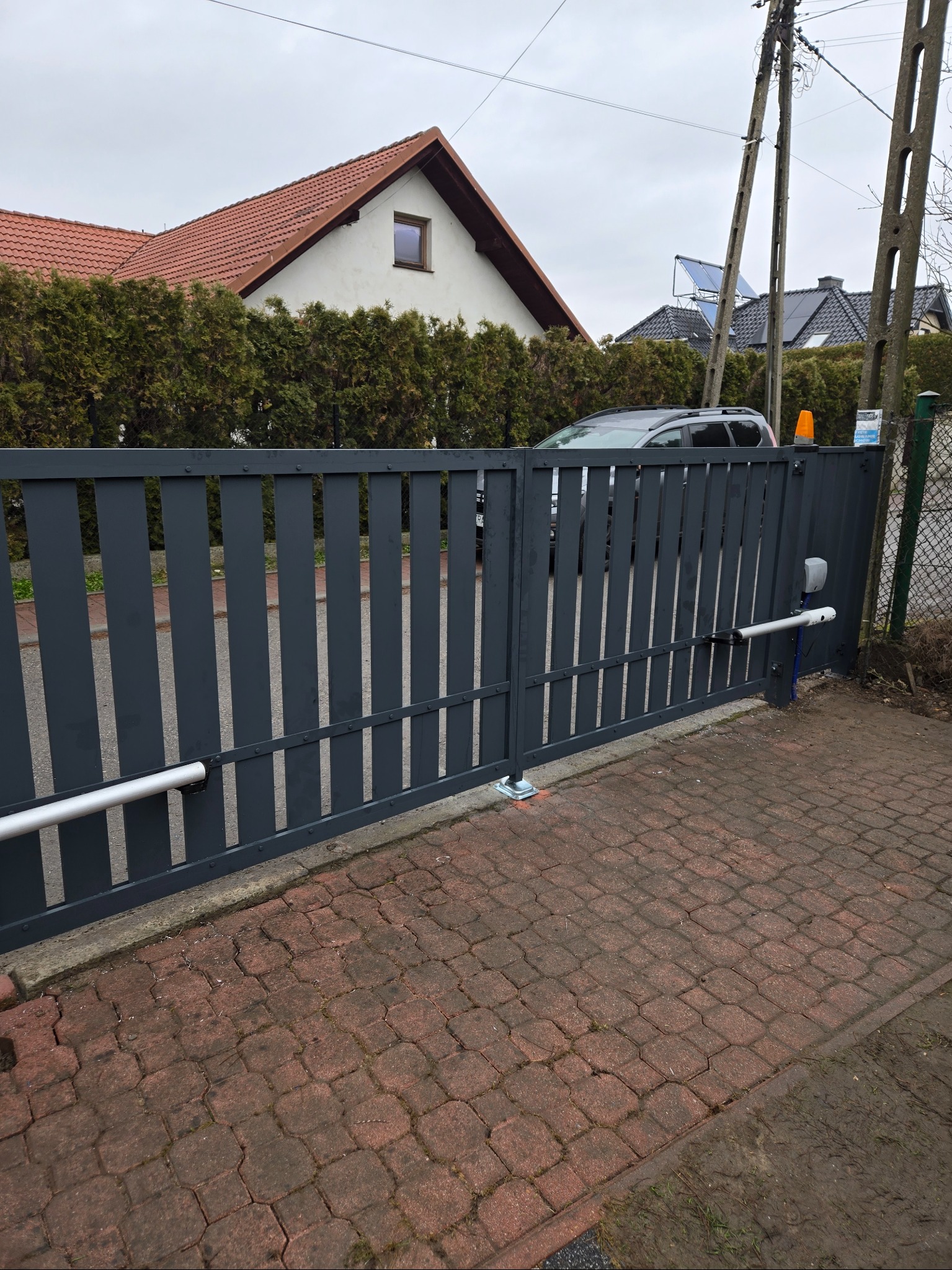 Grafitowa brama wjazdowa z automatyką na posesji z kostki, widok z zewnątrz. Słupki bramy z widocznymi mocowaniami. W tle dom z czerwoną dachówką i zielony żywopłot.