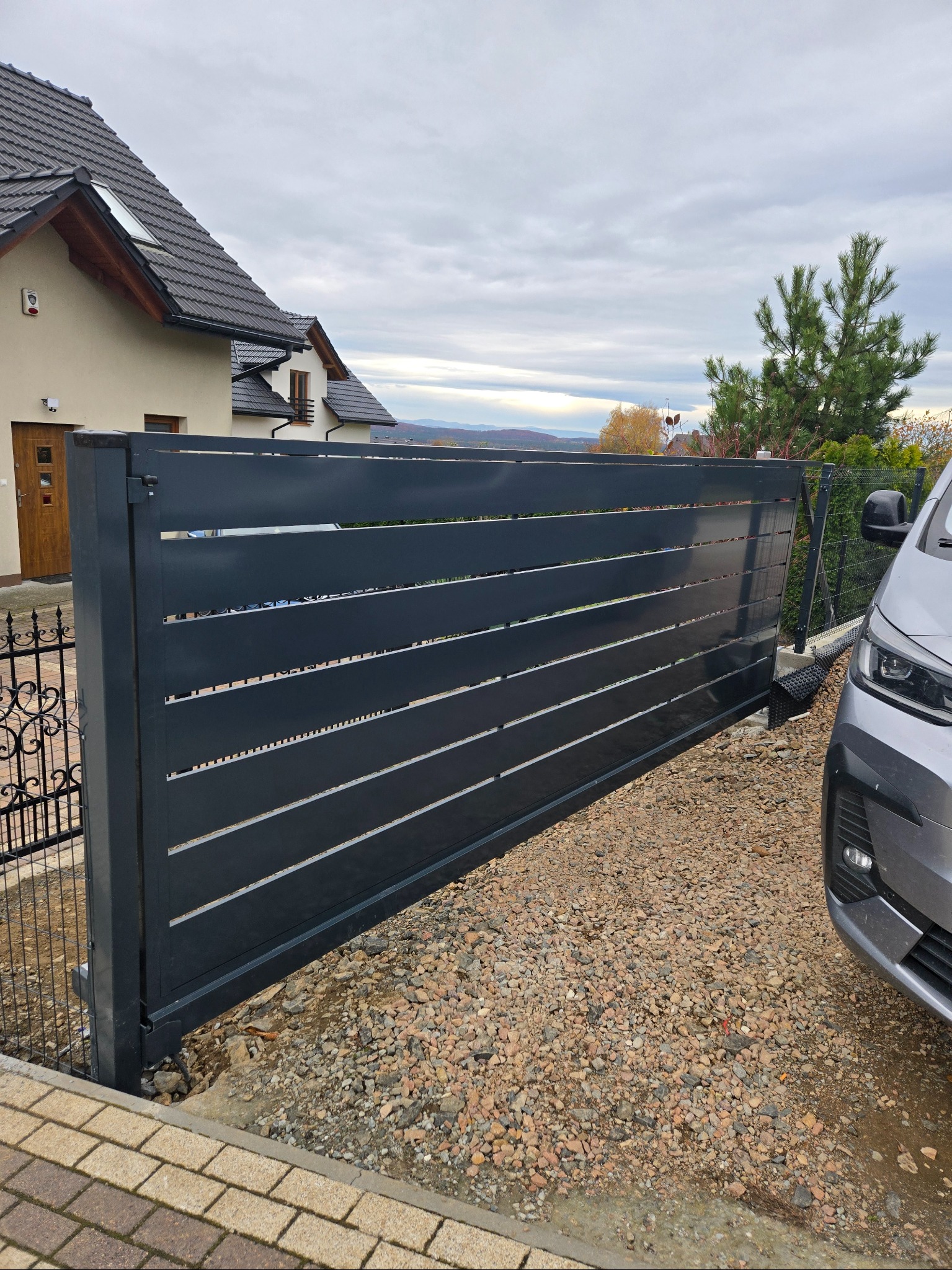 Nowoczesna, przesuwna brama wjazdowa z poziomymi panelami w kolorze antracytowym, częściowo otwarta, na tle domu jednorodzinnego i żwirowego podjazdu.