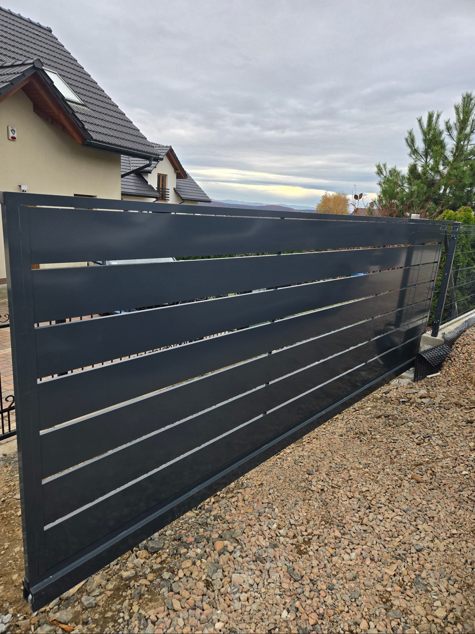 Nowoczesna, przesuwna brama wjazdowa z poziomymi panelami w kolorze antracytowym na tle domu jednorodzinnego i pochmurnego nieba. Widoczne detale konstrukcyjne.