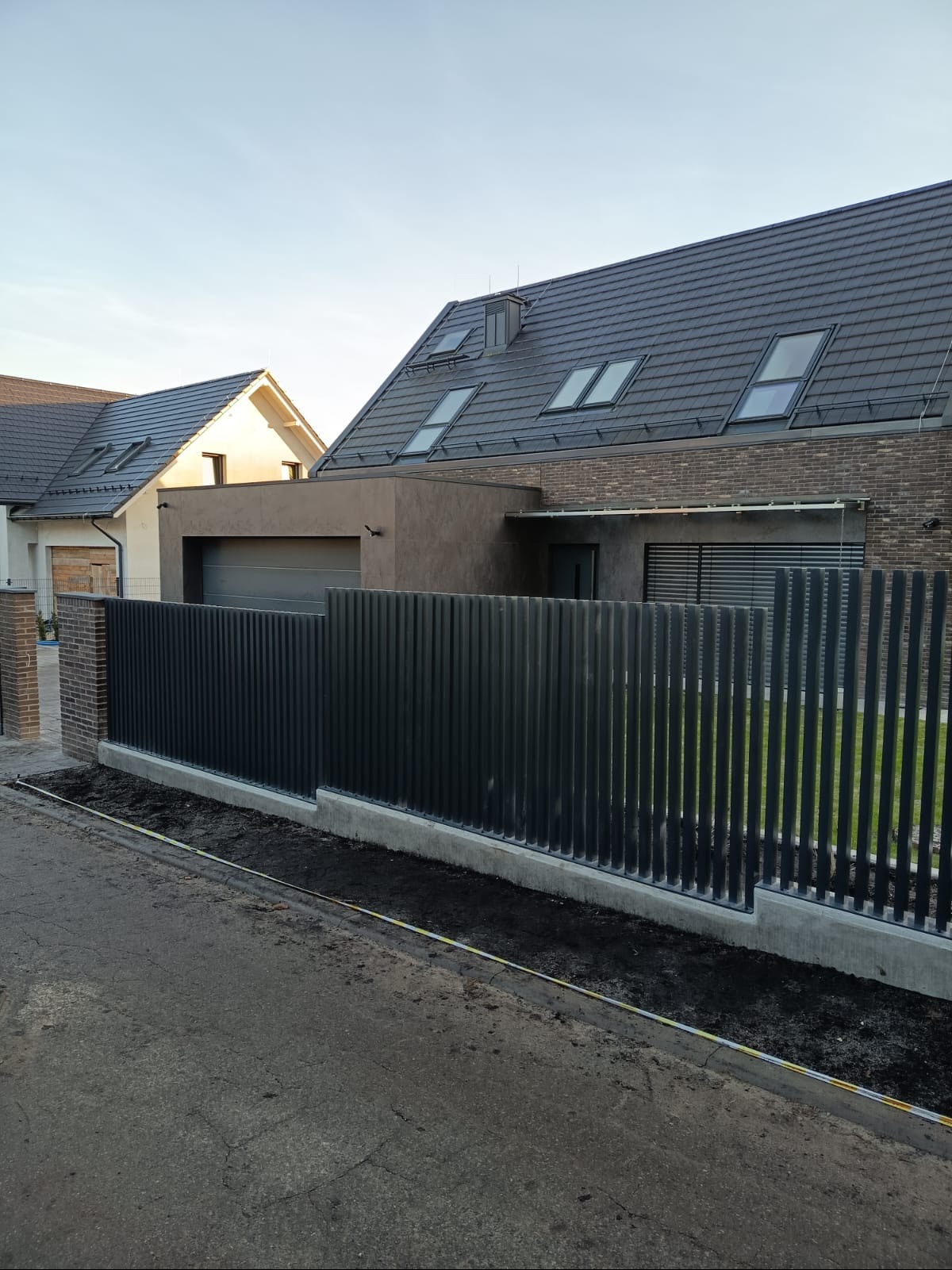 Nowoczesne, pionowe ogrodzenie metalowe w kolorze grafitowym, oddzielające posesję od ulicy. W tle widoczny dom z ciemną dachówką i oknami dachowymi.