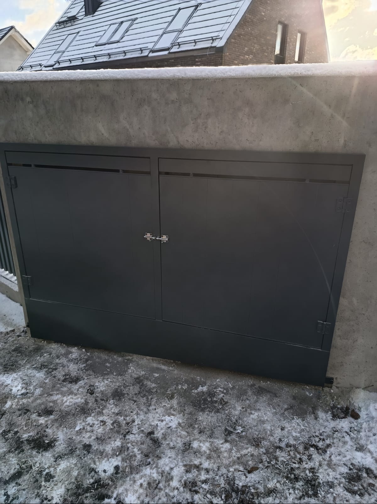 Szara, dwuskrzydłowa brama wjazdowa z metalu, zamontowana w betonowym murze. Zima, śnieg na ziemi. Widoczne zawiasy i zamek. Minimalistyczny design.