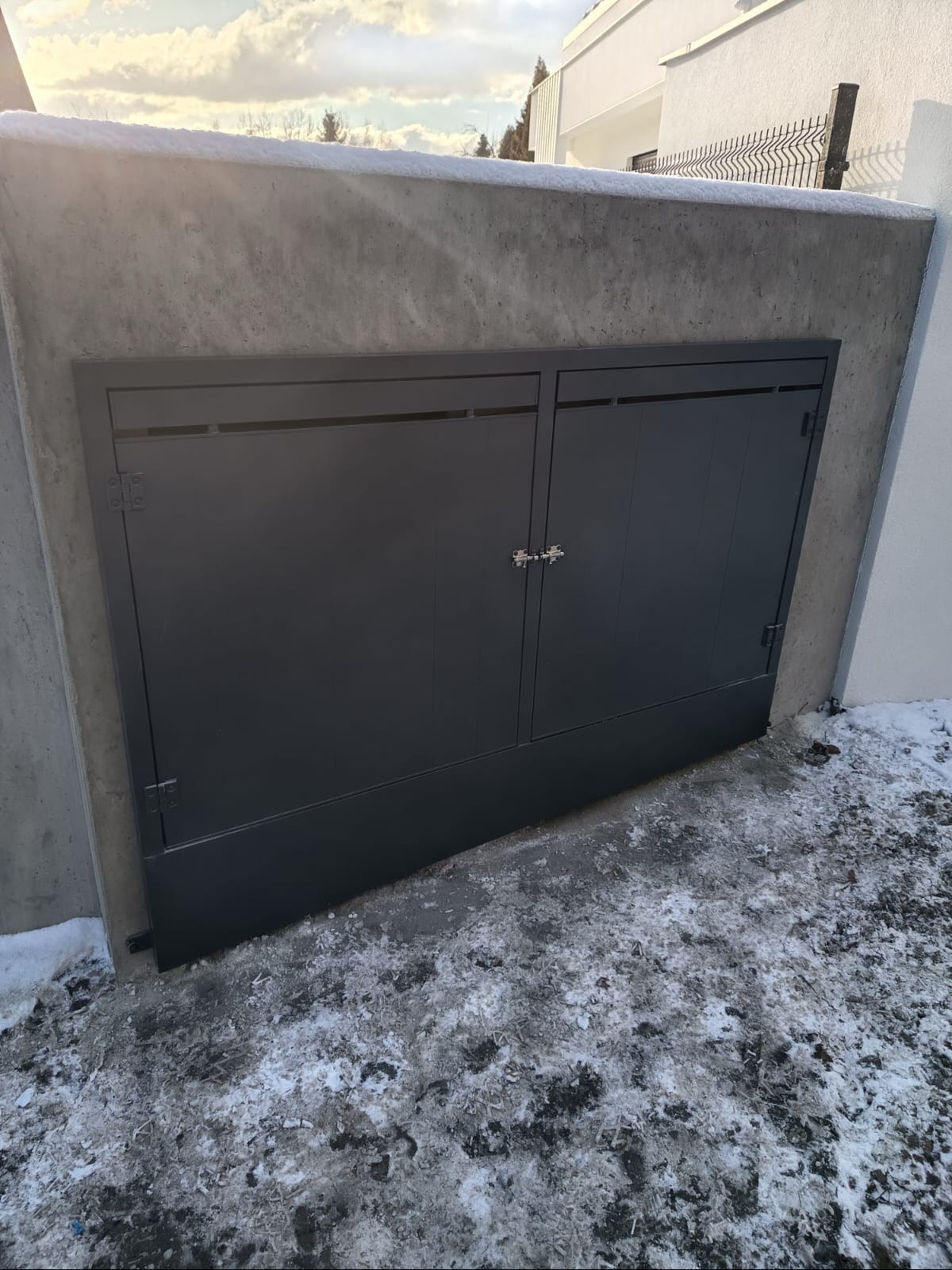 Dwu-skrzydłowa, grafitowa brama garażowa wbudowana w betonowy mur, z widocznymi zawiasami i zamkiem, na tle ośnieżonego podłoża.