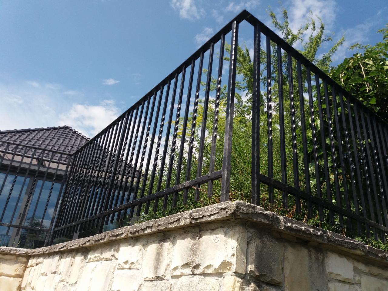 Czarny, metalowy płot z ozdobnymi prętami na kamiennym murku, z zielenią w tle i fragmentem domu z ciemnym dachem.