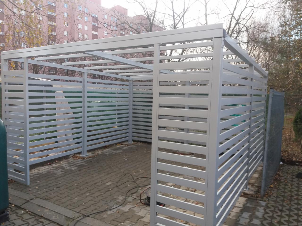 Szara, ażurowa konstrukcja z metalowych profili na kostce brukowej, widoczne przewody elektryczne i budynek w tle.