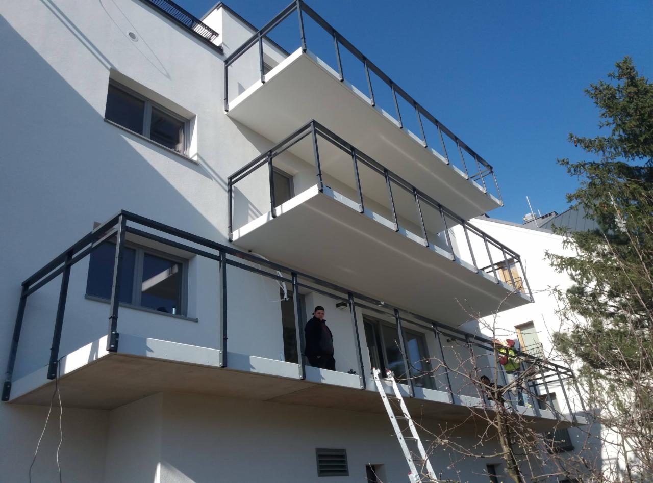 Biały budynek z nowoczesnymi balkonami z czarnymi metalowymi balustradami; jeden pracownik na drabinie, drugi na balkonie w odblaskowej kamizelce.