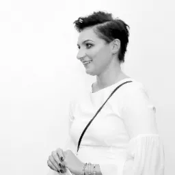 Portret kobiety w białej sukience z paskiem na ramieniu, uśmiechniętej i spoglądającej w bok na jasnym tle.