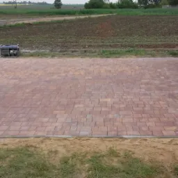 Ułożona kostka brukowa w odcieniach czerwieni i brązu, przylegająca do budynku z brązową bramą garażową, widoczny generator prądu, wózek z kostką i narzędzia w tle.