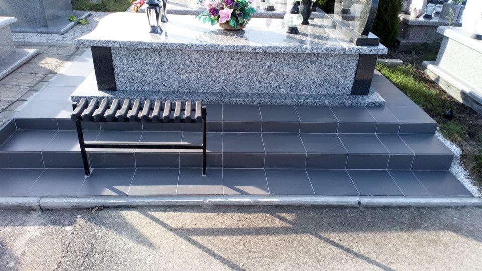 Schody wyłożone szarą, kwadratową płytką prowadzące do granitowego nagrobka z kwiatami i zniczami, z czarną metalową ławeczką z przodu.