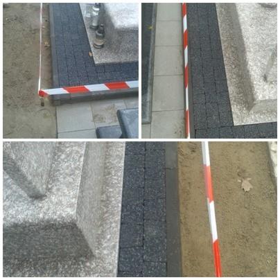 Montaż ciemnej kostki przy nagrobku, oddzielony biało-czerwoną taśmą ostrzegawczą, widoczne fragmenty otoczenia cmentarza.