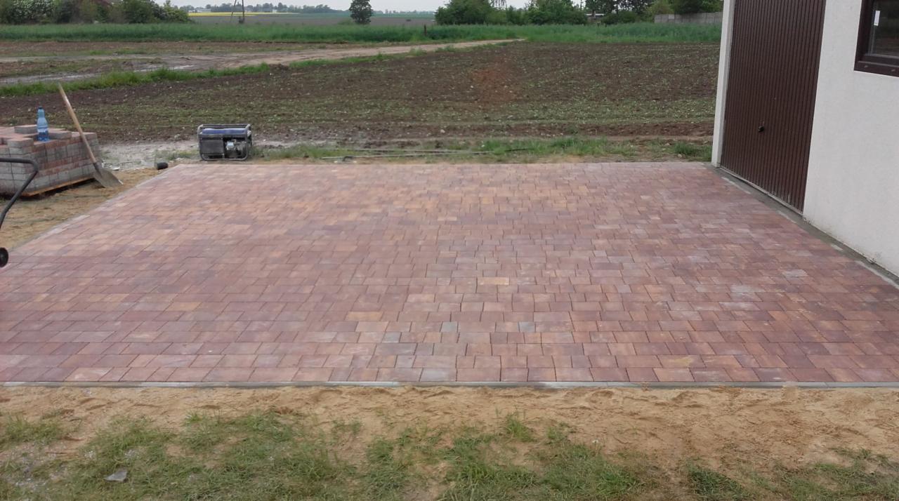 Ułożona kostka brukowa w odcieniach czerwieni i brązu, przylegająca do budynku z brązową bramą garażową, widoczny generator prądu, wózek z kostką i narzędzia w tle.