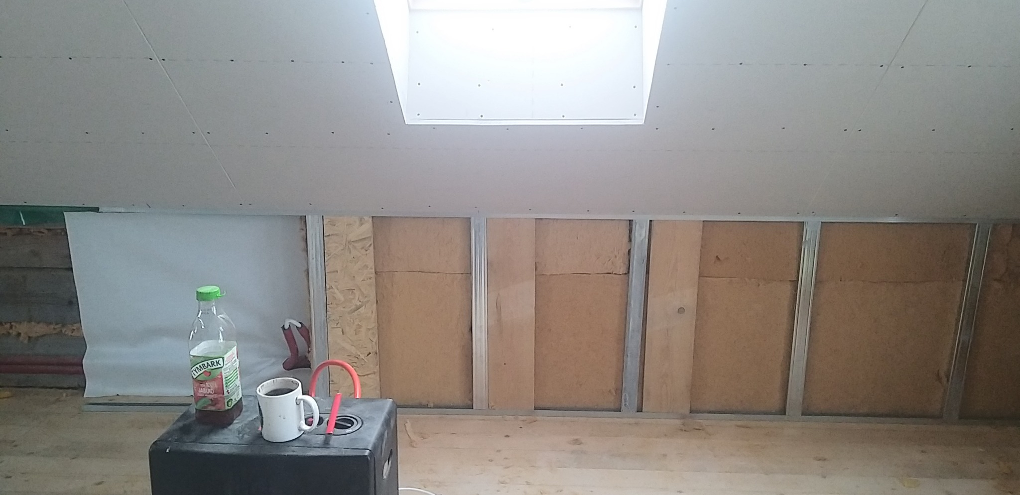 Poddasze w trakcie adaptacji: widoczne ocieplenie wełną mineralną w metalowym stelażu, częściowo zakryte płytą karton-gips. Na pierwszym planie napój i kubek.