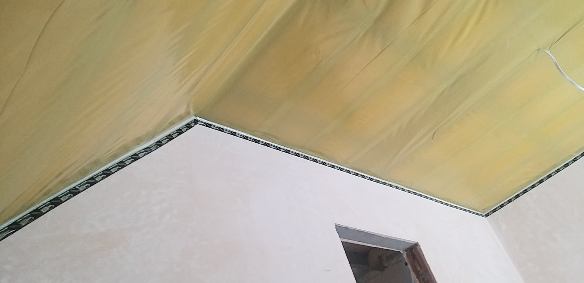 Narożnik pokoju z folią malarską na suficie, zabezpieczającą przed zabrudzeniem. Ściany po szpachlowaniu, przygotowane do malowania. Widoczny fragment ościeżnicy.