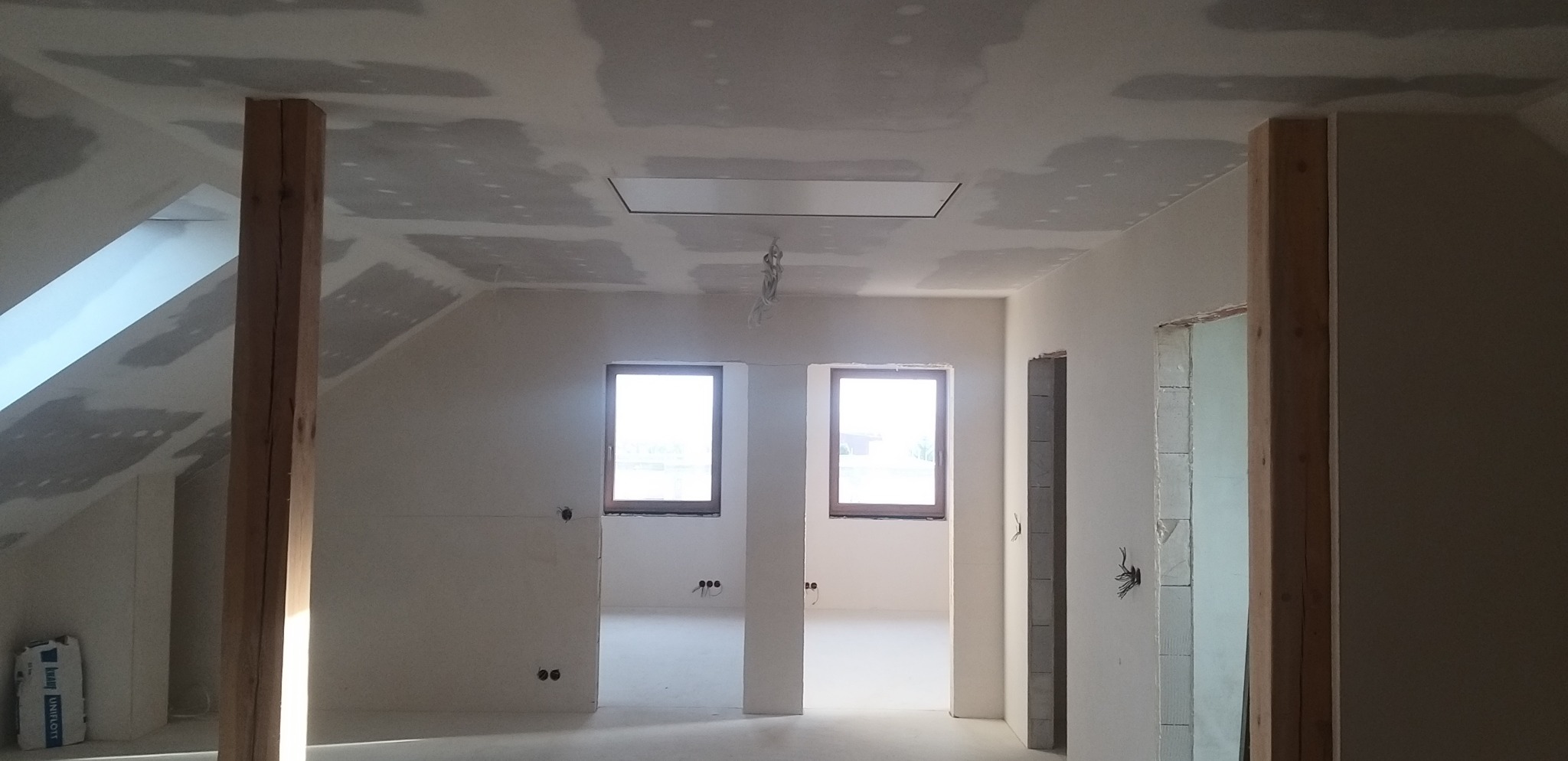 Poddasze w trakcie prac wykończeniowych: ściany i sufit pokryte gładzią, widoczne drewniane słupy konstrukcyjne, okna z naturalnym światłem. Etap przygotowania do malowania.