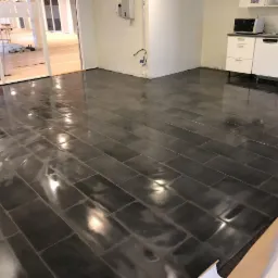 Świeżo ułożona, błyszcząca, ciemnoszara podłoga z płytek w kształcie prostokątów, widoczne odblaski światła, fragment kuchni z białymi szafkami w tle, dalsza część pomieszczenia z przeszkloną ścianą.