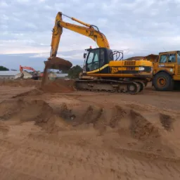 Żółto-czarna koparka JCB nabiera ziemię na placu budowy, w tle widoczny wywrotka i druga koparka, pochmurne niebo.