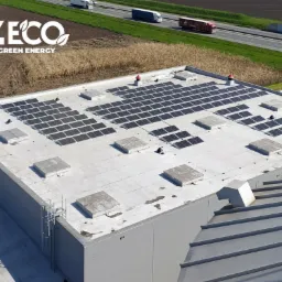 Widok z góry na dach przemysłowego budynku z zamontowanymi panelami fotowoltaicznymi; w tle droga z jadącymi samochodami ciężarowymi i pole uprawne; widoczne logo firmy.