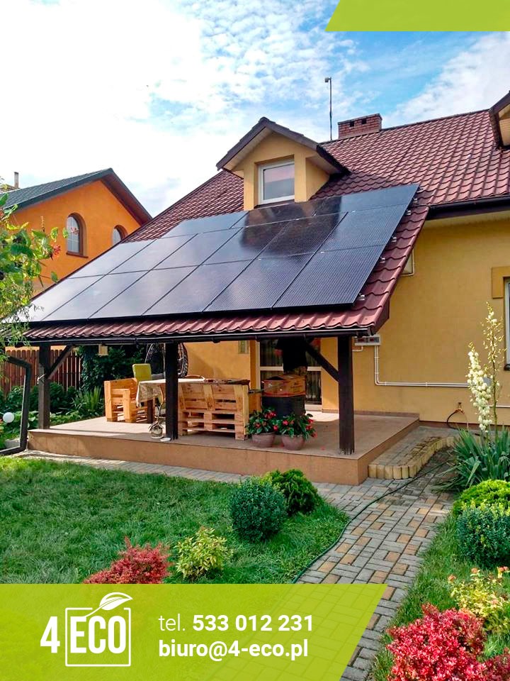 Dom jednorodzinny z panelami słonecznymi na dachu, widok z ogrodu z drewnianym tarasem i meblami z palet, w tle błękitne niebo z chmurami.