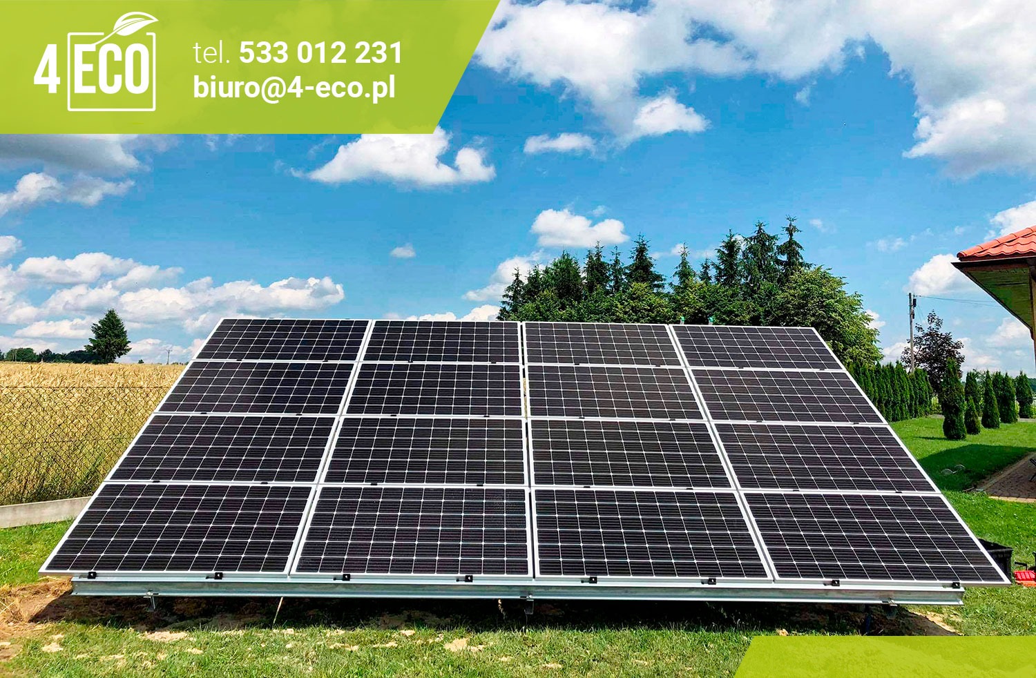 Instalacja paneli słonecznych na metalowej konstrukcji, widok z niskiej perspektywy, na tle błękitnego nieba z chmurami i zieleni drzew oraz części dachu budynku.