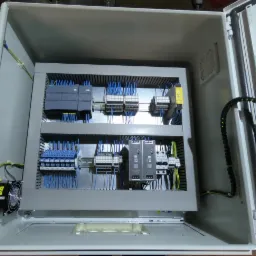 Otwarta szafa sterownicza z modułami PLC Siemens, bezpiecznikami, przekaźnikami i okablowaniem w kolorze niebieskim, widoczny wentylator chłodzący.