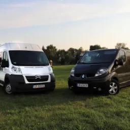 Dwa busy: biały Citroen i czarny Renault, stojące na trawiastym terenie, w tle widoczna wieża kościoła.