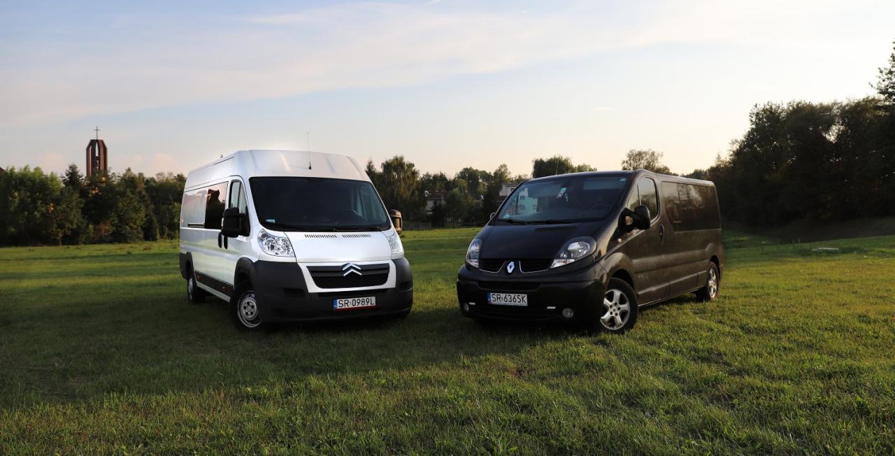 Dwa busy: biały Citroen i czarny Renault, stojące na trawiastym terenie, w tle widoczna wieża kościoła.