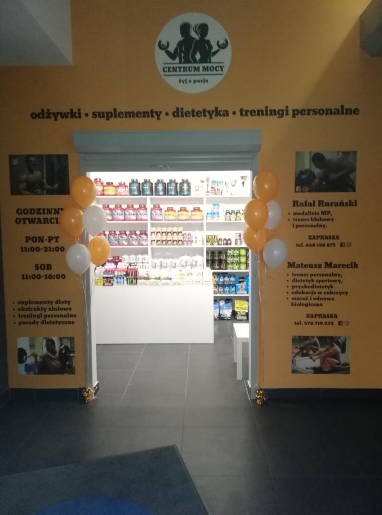 Wejście do Centrum Mocy z ofertą odżywek, suplementów, dietetyki i treningów personalnych, udekorowane białymi i pomarańczowymi balonami.