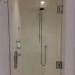 Nowoczesna kabina prysznicowa z deszczownicą i szklanymi drzwiami na zawiasach, widoczny odpływ liniowy w podłodze wyłożonej białym marmurem, logo firmy w prawym dolnym rogu.