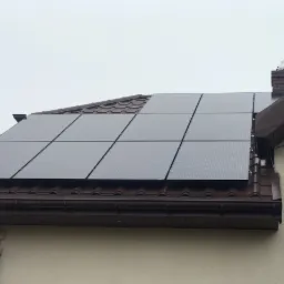 Panele słoneczne na brązowym dachu domu jednorodzinnego, widoczny komin z atrapą wiatrowskazu, elewacja z elementami kamiennymi.