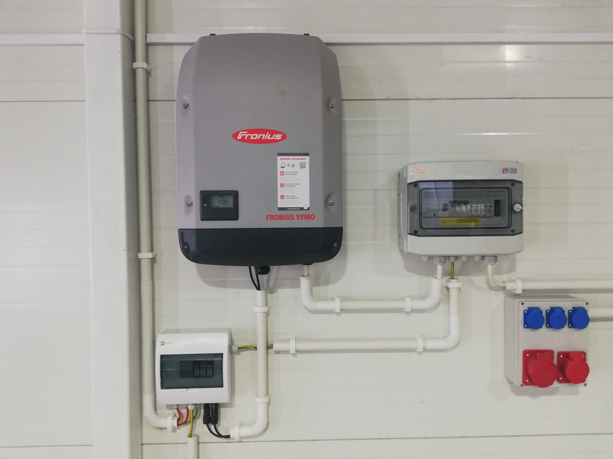 Szary falownik Fronius Symo z podłączonymi przewodami i skrzynkami elektrycznymi na białej ścianie z paneli. Widoczne zabezpieczenia i gniazda elektryczne.