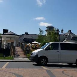 Biały van zaparkowany przed restauracją z mostkiem i wieżą w tle, słoneczny dzień.