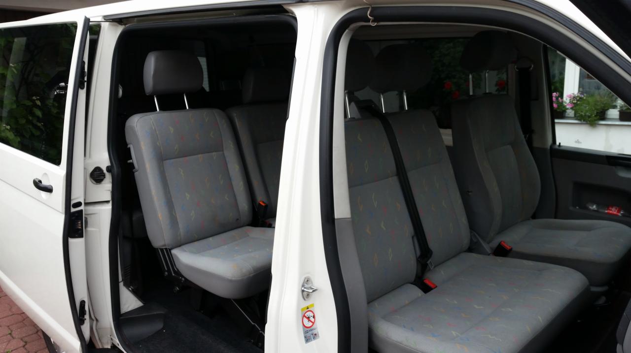 Wnętrze białego busa z otwartymi drzwiami, widoczne szare fotele z wzorem i zagłówkami, z zewnątrz za oknem widać fragment zieleni i kwiaty w doniczkach na parapecie.