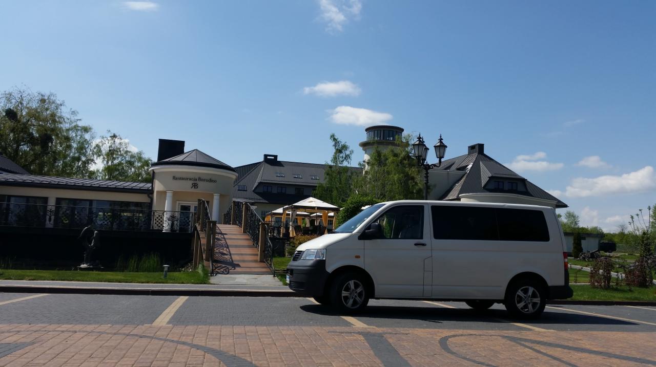 Biały van zaparkowany przed restauracją z mostkiem i wieżą w tle, słoneczny dzień.