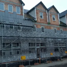 Budynek wielorodzinny w trakcie ocieplania elewacji, widoczne rusztowania i płyty izolacyjne z białymi kołkami mocującymi.